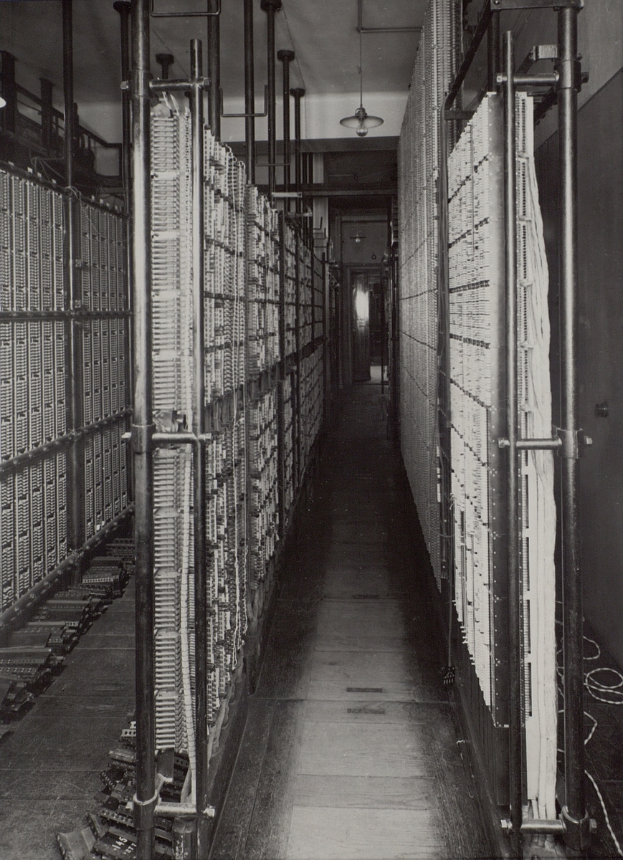 Ein Schwarz-Weiß-Foto eines langen Ganges in einem Rechenzentrum, das Metallobjekte, Rohre und Deckenleuchten sowie zahlreiche Server und mit Daten gefüllte Gestelle zeigt.