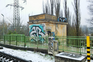 Eine Bahnstrecke mit Graffiti an der Seite, umgeben von Gelöndern, Pfosten, Lichtern und Bäumen, mit Törmen und Drähten im Hintergrund und Schnee am Boden.