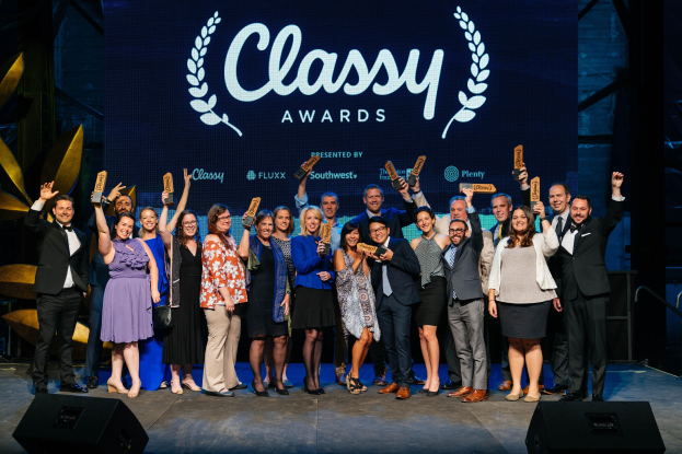 Gruppe von Menschen auf einer Bühne mit Auszeichnungen, Lautsprechern darunter und einem Bildschirm mit der Aufschrift "Classy Awards 2019" im Hintergrund.