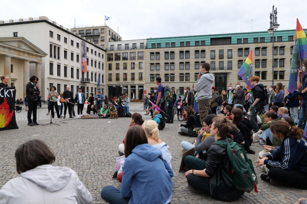 Gruppe von Menschen, die auf dem Boden vor einer Menge mit Fahnen und Spruchbändern sitzen, eine Person spricht in ein Mikrofon, eine Statue auf einem Podest und Gebäude im Hintergrund bei einer Demonstration gegen Homosexuelle in Berlin, Deutschland.