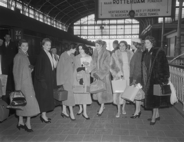 Schwarz-weißes Bild einer Gruppe von Frauen, die an einem Bahnhof stehen und Taschen halten, mit einem Zug links und Geländern und einer Tafel rechts.