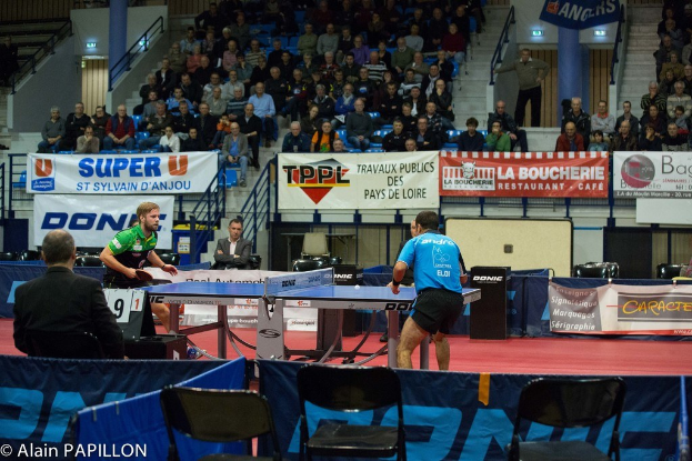 Indoor-Tischtennis-Match mit spielenden Spielern, sitzenden und stehenden Zuschauern, leeren Stühlen und Werbeplakaten im Hintergrund.