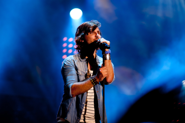 Ein Mann steht auf einer Bühne unter hellen Scheinwerfern und singt in ein Mikrofon, trägt ein schwarzes T-Shirt und Jeans mit einer Gitarre, stylishe Haare und einen selbstbewussten Ausdruck.