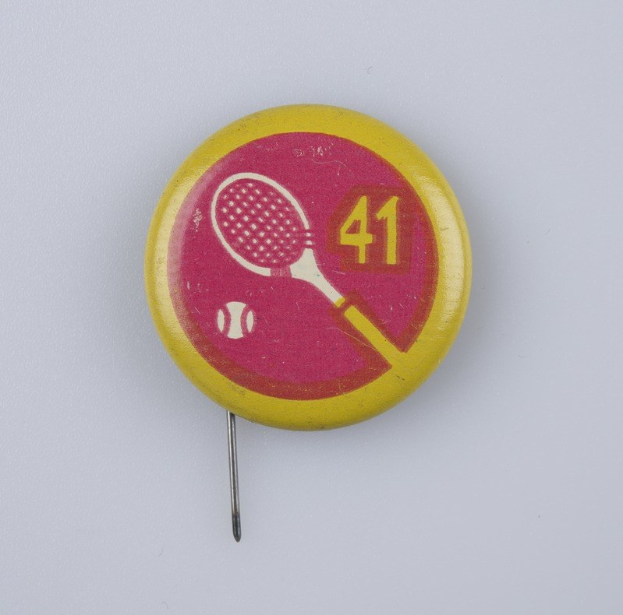 Gelb-rosa Abzeichen mit einem Tennisracket-Design, mit Text und Zahlen, auf einer weißen Oberfläche angeheftet.
