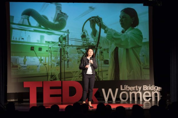 Frau auf einer Bühne mit Mikrofon, dahinter ein Display mit Bild einer Frau und Text zum TEDxWomen-Event.