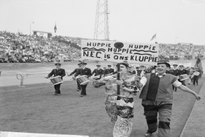 Eine Gruppe von Menschen, die draußen spazieren gehen, einige halten Musikinstrumente und ein Schild mit der Aufschrift "Huppie Huppie NEC ist auf Kluppie", mit einem Metallzaun, einem Turm, Polen, Flaggen, einem Gebäude und einem bewölkten Himmel im Hintergrund.