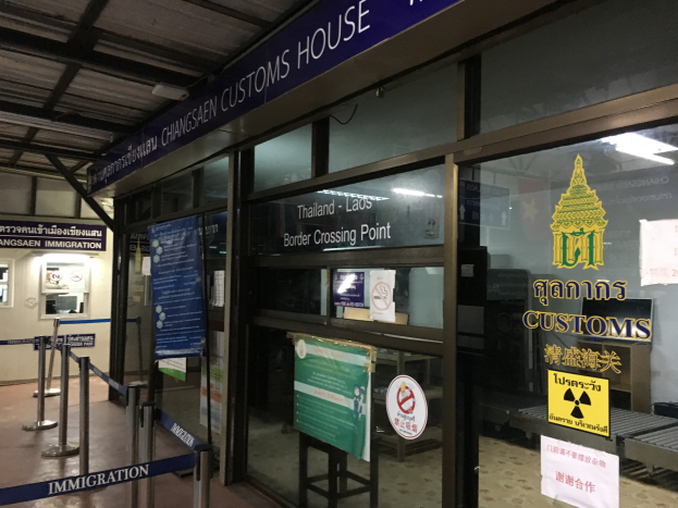 Der Eingang eines Zollhauses in Bangkok, Thailand, mit Glaswänden und -türen, Schildern mit Text und Bildern, Pfosten mit Bändern, einem Schild, einem Dach mit Deckenleuchten und einer Wand mit Gegenständen.