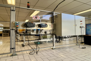 Große industrielle Automationsmaschine in einer Einrichtung, mit einer Steuerkonsole auf der rechten Seite, Deckenleuchten und einer Wand im Hintergrund.