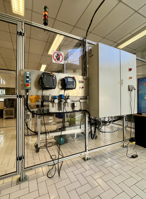 Große industrielle Automationsmaschine in einer Einrichtung, mit einer Steuerkonsole auf der rechten Seite, Deckenleuchten und einer Wand im Hintergrund.