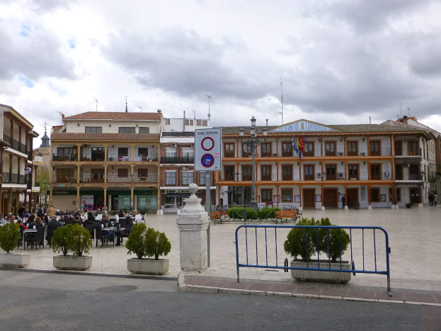 Ein belebter Stadtplatz mit Menschen, die sitzen und stehen, Topfpflanzen, eine Metallabsperrung, ein Schild an einem Pfahl, Straßenlaternen mit Flaggen, umliegende Gebäude und ein bewölkter Himmel.