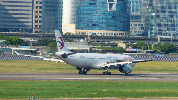 Großflugzeug beim Start von einer Flughafenpiste, umgeben von Gras, einem Zaun, Bäumen, Straßenlaternen, Schildern, einer Brücke, Gebäuden mit Fenstern und einem bewölkten Himmel.