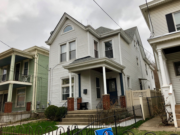 Reihe von Häusern mit Fenstern, Türen, Säulen, Geländern, Treppen und Dächern, umgeben von einem Metallzaun, Gras, Pflanzen, Bäumen und einem bewölkten Himmel, mit einer Mietanzeige am unteren Rand, die die Adresse und die Listen-details anzeigt.