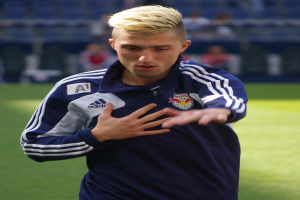 Ein junger Mann in einem blauen T-Shirt steht auf einem Fußballfeld und streckt die Arme aus, sein unscharfer Hintergrund suggeriert ein Fußballspiel.