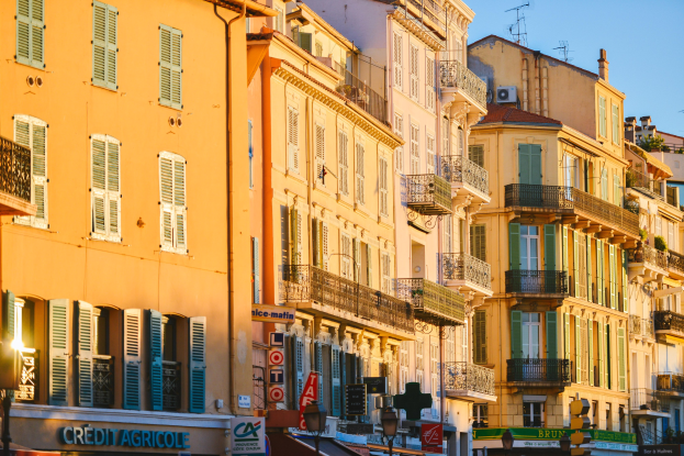 Eine Stadtstraße mit hohen Gebäuden, die Fenster, Geländer und Balkone sowie Straßenschilder, Pfosten, Laternen, Verkehrssignale, Bäume und einen klaren blauen Himmel zeigen.