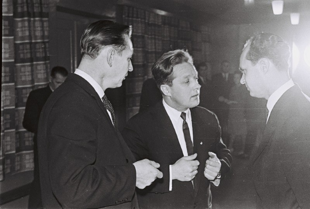 Drei Männer in Anzügen unterhalten sich in einem schwach beleuchteten Raum mit beleuchteten Bücherregalen im Hintergrund.