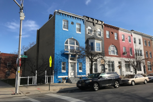 Eine Reihe bunter Häuser an einer Straßenecke in Baltimore, Maryland, mit parkenden Autos, Bäumen, Pfählen, Brettern und einem Zaun, der die Straße säumt.