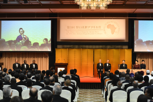 Eine Gruppe von Menschen sitzt in einem Raum mit einem Podium, Mikrofonen und einem großen Bildschirm, der Text und Bilder anzeigt, sowie einer mit Kristallleuchtern geschmückten Decke darüber.