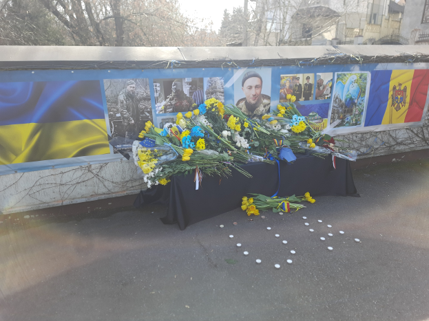 Eine improvisierte Gedenkstätte in Kiew, Ukraine, mit Blumensträußen auf der Straße vor einer Wand mit Fotos und Flaggen, vor einer Kulisse aus Bäumen, Gebäuden und einem klaren blauen Himmel.