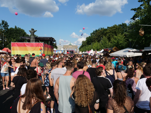 Eine große Menge geht eine Straße mit Zelten, Bäumen, Masten, Lichtern und einer Statue entlang, mit Gebäuden und einem bewölkten Himmel mit Ballons im Hintergrund beim Berlin Pride Festival.