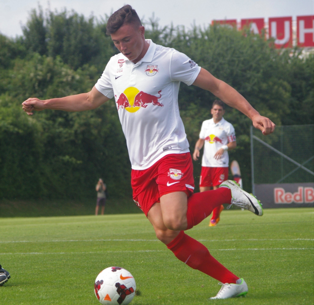 Ein professioneller Fußballspieler in roter Uniform kickt einen Fußball auf einem grünen Feld mit Bäumen und einem klaren blauen Himmel im Hintergrund, mit "RB Leipzig" auf einer Tafel.