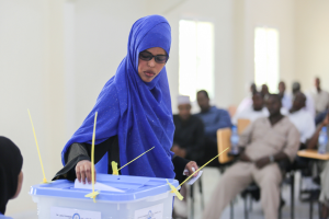 Eine Frau mit blauem Kopftuch gibt ihre Stimme in einer Wahllokal ab, umgeben von Menschen auf Stühlen und einer fensterumrahmten Wand im Hintergrund.