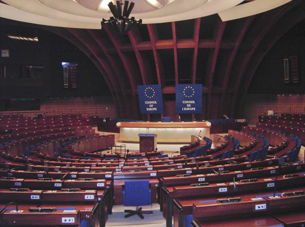 Großer halbrunder Konferenzraum des Europäischen Parlaments in Brüssel mit Stufenbestuhlung, zentralem Podium, wandmontierten Texttafeln und prunkvollem Deckenlicht.