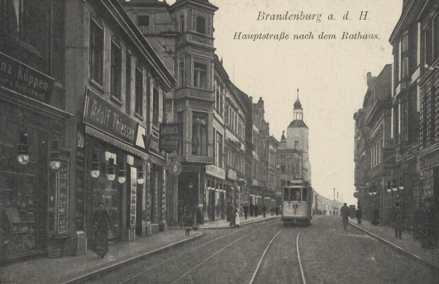 Schwarzes und weißes Foto einer Stadtstraße in Brandenburg, Deutschland mit einer Tram, Fußgängern, Gebäuden, Schildern, Straßenlaternen und einem bewölkten Himmel.