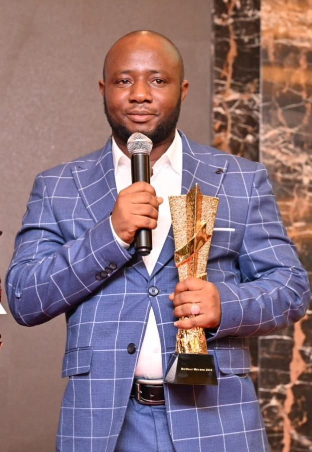 Ein Mann in einem blauen Anzug hält ein Mikrofon und eine Trophäe und steht selbstbewusst mit einem freudigen Gesichtsausdruck da, was darauf hindeutet, dass er gerade einen Preis gewonnen hat.