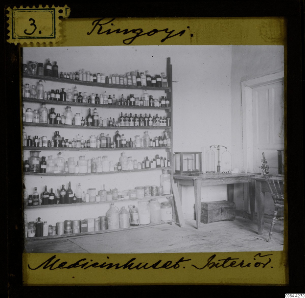 Schwarzes und weißes Foto eines alten Apothekeninneren mit Regalen voller Flaschen, einem Tisch mit Gegenständen, einem Stuhl, einer Tür und einer Wand, mit Text unten.