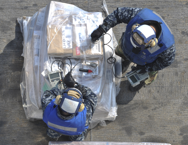 Zwei Männer in blauen Uniformen und Helmen arbeiten an einem großen Drohnenmodell und halten verschiedene Gegenstände mit einer Plastikhülle auf dem Boden.