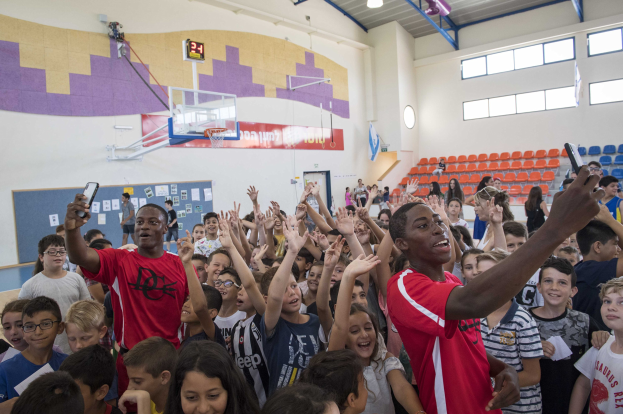 Kinder vor einem Basketballplatz mit Handys während eines Basketballcamps, mit einem Tor, einem Basketballkorb, einer Uhr, angeklebten Papieren an einer Tafel, Deckenlampen, Stühlen und Fenstern im Hintergrund.