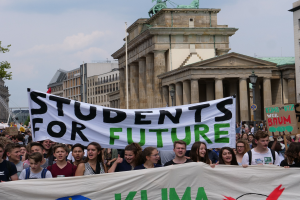 Eine Gruppe von Schülern marschiert in Berlin, die ein buntes "Students for Future"-Plakat trägt, vor einem Hintergrund aus Gebäuden, Bäumen und Himmel.