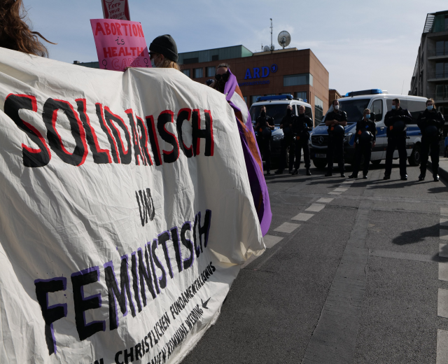 Gruppe von Menschen marschiert die Straße entlang und hölt eine 'Solidarität und Feminismus'-Fahne, mit geparkten Fahrzeugen, Gebäuden, einer Schüsselantenne und einem klaren blauen Himmel im Hintergrund.