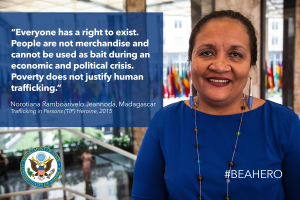 Eine Frau in einem blauen Kleid steht lachend vor einem Gebäude mit einem Anti-Menschenhandels-Spruch, mit Menschen, Pflanzen und Flaggen im Hintergrund.