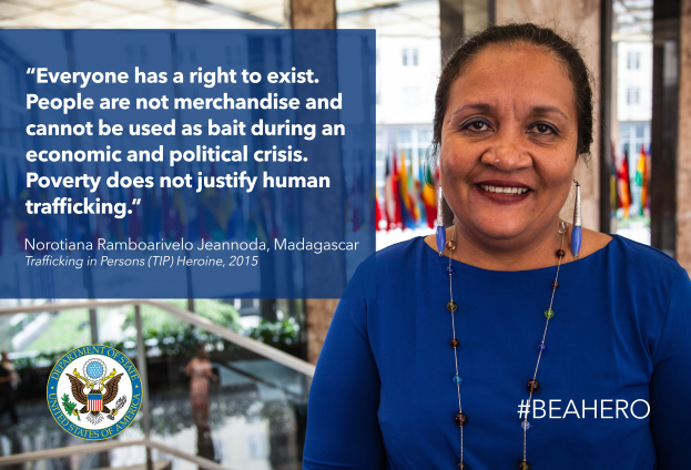 Eine Frau in einem blauen Kleid steht lachend vor einem Gebäude mit einem Anti-Menschenhandels-Spruch, mit Menschen, Pflanzen und Flaggen im Hintergrund.