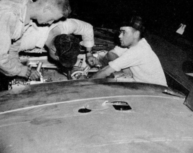 Drei Männer in Mützen arbeiten an einem schwarz-weißen Auto in einer Garage, umgeben von Werkzeugen und Reparaturmaterialien.