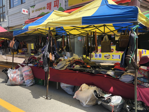 Ein Straßenmarkt mit Zelten, die Taschen, Hüte und andere Artikel präsentieren, vor einem Hintergrund aus Gebäuden mit Fenstern und Texttafeln.