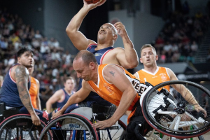Eine Gruppe von Männern in Rollstühlen beim Basketballspielen, einer hält den Ball, mit Zuschauern im Hintergrund.