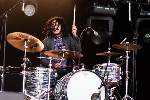 Ein Mann mit Dreadlocks, der auf der Bühne Schlagzeug spielt, hält Drumsticks in den Händen und hat Mikrofonständer vor sich. Im Hintergrund sind Scheinwerfer und Equipment in einer dunklen Atmosphäre zu sehen.