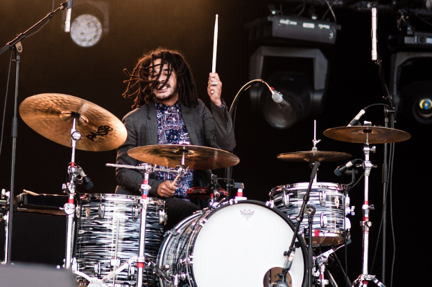 Ein Mann mit Dreadlocks, der auf der Bühne Schlagzeug spielt, hält Drumsticks in den Händen und hat Mikrofonständer vor sich. Im Hintergrund sind Scheinwerfer und Equipment in einer dunklen Atmosphäre zu sehen.