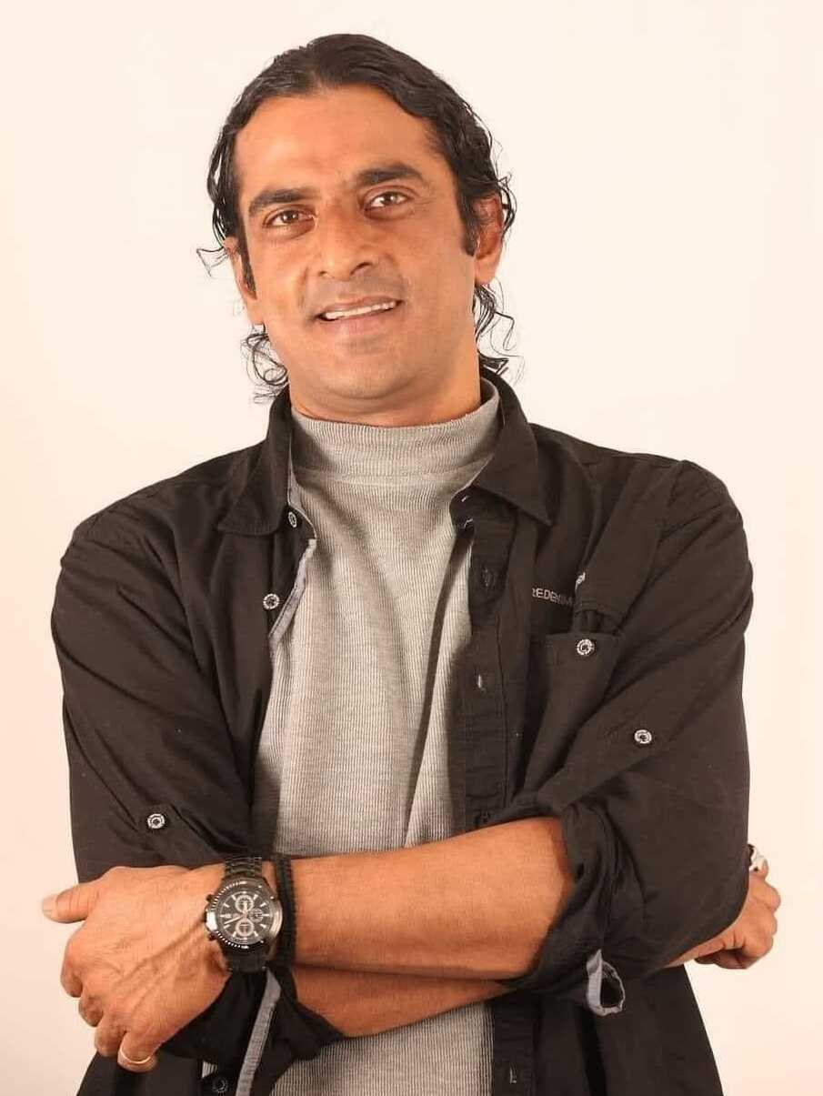 Ein Mann in einem schwarzen und aschfarbenen Kleid, der eine Uhr tr├Ągt, steht mit überkreuzten Armen vor einem wei├čen Hintergrund und sieht nachdenklich aus.