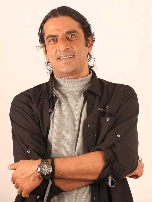 Ein Mann in einem schwarzen und aschfarbenen Kleid, der eine Uhr tr├Ągt, steht mit überkreuzten Armen vor einem wei├čen Hintergrund und sieht nachdenklich aus.