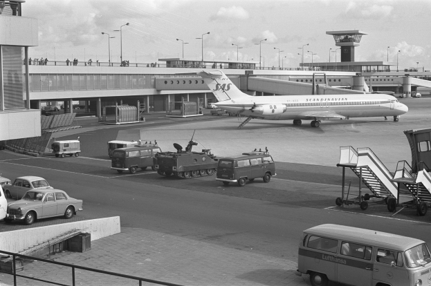Ein Schwarz-Weiß-Foto eines Flughafens mit Autos und Lastwagen auf der Straße, einem Flugzeug auf der Rollbahn, Treppen mit Geländern, einem Gebäude mit Säulen und Geländern im Hintergrund, Laternenmasten und einem bewölkten Himmel.