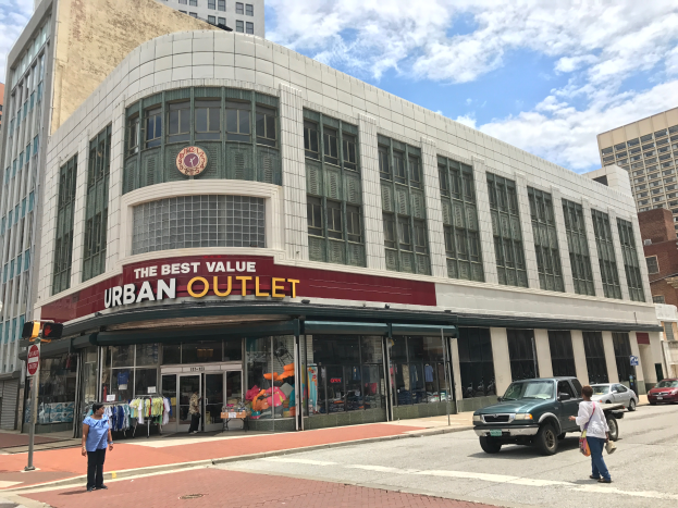 Eine belebte Stadtstraße mit Fahrzeugen, Fußgängern, hohen Gebäuden und einem Schild mit der Aufschrift "The Best Value Urban Outlet" unter einem bewölkten Himmel.