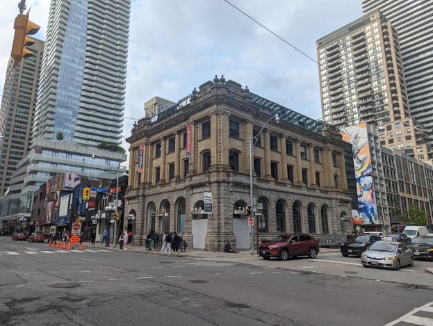 Eine belebte Stadtstraße mit hohen Gebäuden im Hintergrund, Fahrzeugen, Fußgängern, Verkehrskegeln, Laternen, Verkehrsampeln, Tafeln, Werbetafeln, Bäumen und der Toronto Public Library als Mittelpunkt unter einem bewölkten Himmel.