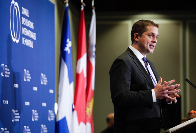 Der kanadische Premierminister in Anzug und Krawatte bei einer Rede an einem Rednerpult mit Mikrofonen, umgeben von Fahnen und einem Banner, mit ein paar Menschen im Hintergrund.