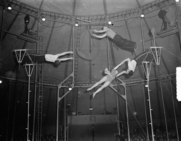 Ein Schwarz-Weiß-Foto von Artisten, die in einem Zirkuszelt Akrobatik zeigen, mit Stangen, Leitern und Lichtern, während die Menge unten zusieht.