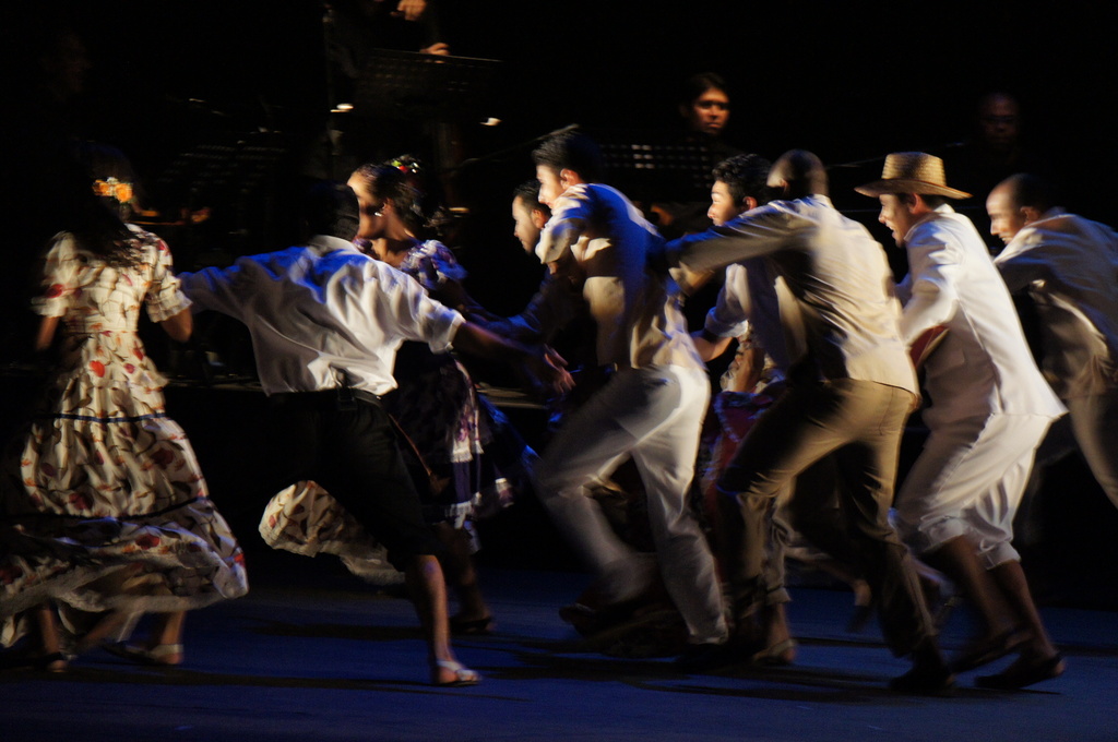 Eine Gruppe von Menschen tanzt im Vordergrund mit einer anderen Gruppe, Ständen und Mikrofonen im Hintergrund, alles auf einem Boden.