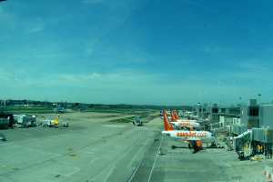 Eine Flughafen-Szene mit Flugzeugen, Gebäuden, Masten, Fahrzeugen und Bäumen, mit einem Himmel darüber und einer Straße darunter und einem Zaun im Vordergrund.