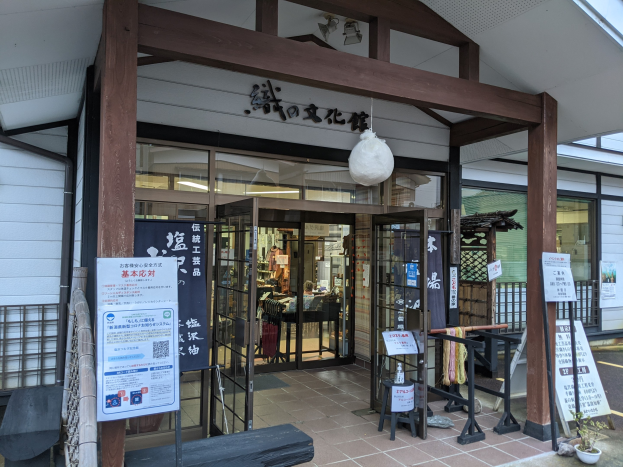 Eingang zu einem japanischen Restaurant mit Glaswänden und -türen, mit einer Tafel, einer Bank, einem Topf mit einer Pflanze, Pfosten, einem Metallzaun, Deckenleuchten und Innenraumtischgestaltung.